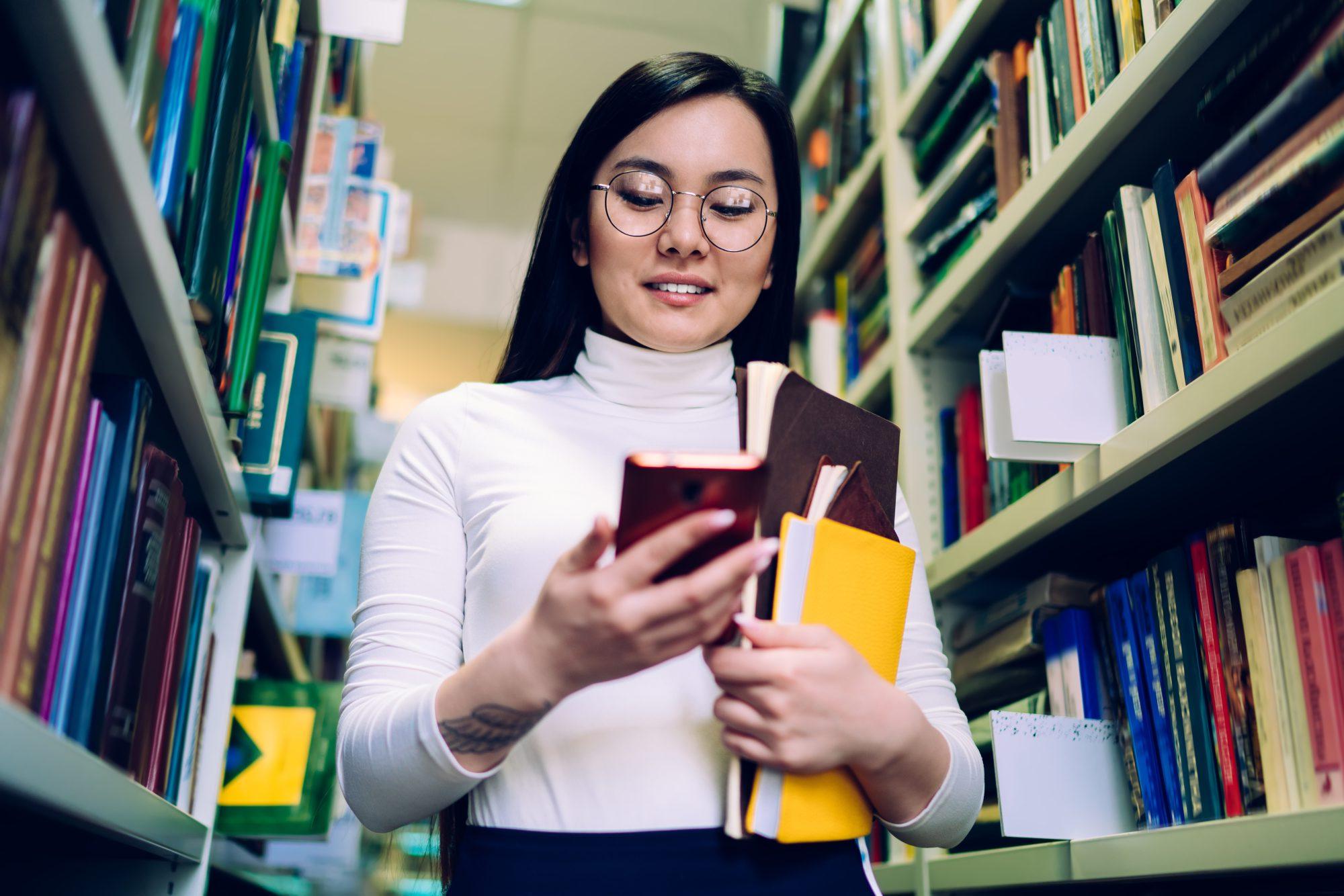 Smiling black haired young Asian female in white top and glasses with books using cellphone standing among rows of bookshelves in library